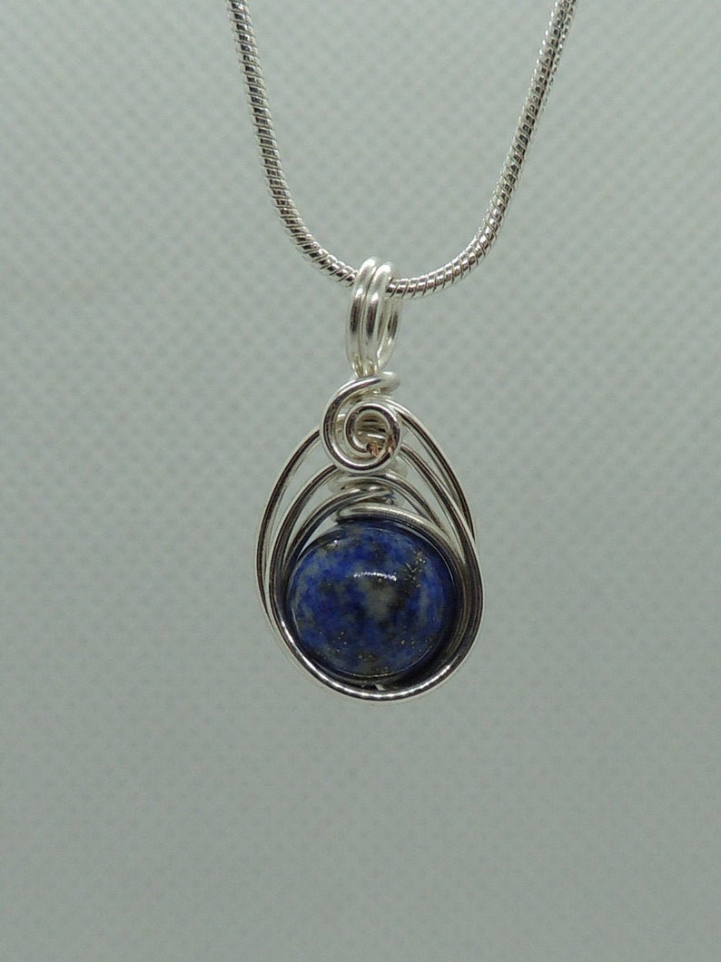 Puede incluir: Un colgante de alambre plateado que envuelve una piedra preciosa redonda de lapisl&aacute;zuli azul profundo con sutiles motas doradas. El colgante cuelga de una cadena de serpiente plateada a juego con una argolla segura en la parte superior.