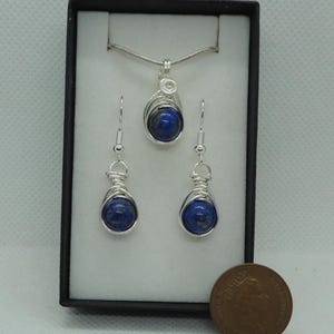 Puede incluir: Un conjunto de joyer&iacute;a que consta de un colgante en una cadena plateada y dos pendientes a juego. Cada pieza presenta una piedra redonda de color azul intenso envuelta en alambre plateado, presentado en una caja negra junto a una moneda.