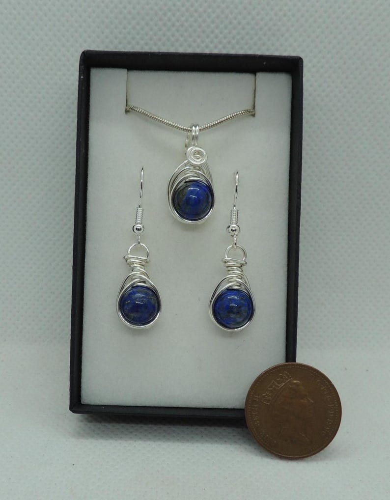 Puede incluir: Un conjunto de joyer&iacute;a que consta de un colgante en una cadena plateada y dos pendientes a juego. Cada pieza presenta una piedra redonda de color azul intenso envuelta en alambre plateado, presentado en una caja negra junto a una moneda.