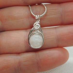 Puede incluir: Un colgante de alambre plateado que envuelve una piedra redonda de color blanco opaco. El alambre de metal forma una jaula en forma de l&aacute;grima alrededor de la piedra con un detalle en espiral en la parte superior. El colgante cuelga de una cadena fina.