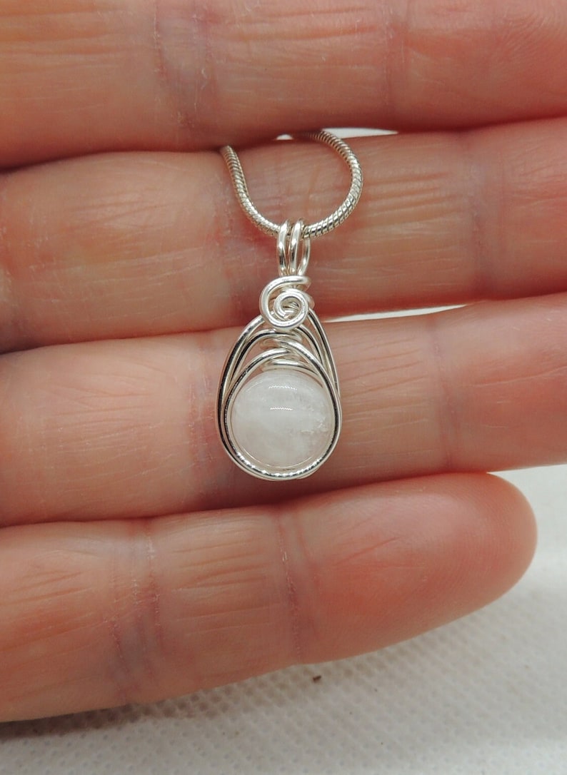 Puede incluir: Un colgante de alambre plateado que envuelve una piedra redonda de color blanco opaco. El alambre de metal forma una jaula en forma de l&aacute;grima alrededor de la piedra con un detalle en espiral en la parte superior. El colgante cuelga de una cadena fina.