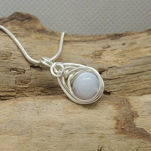 May include: A silver-toned wire-wrapped pendant featuring a smooth, round, pale blue stone. The pendant is suspended from a matching silver-toned snake chain, with the wire coiled in a teardrop shape around the central gemstone setting.