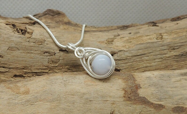 May include: A silver-toned wire-wrapped pendant featuring a smooth, round, pale blue stone. The pendant is suspended from a matching silver-toned snake chain, with the wire coiled in a teardrop shape around the central gemstone setting.