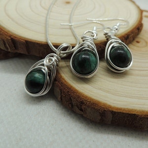 Puede incluir: Un conjunto de joyer&iacute;a que consta de un colgante en una cadena de tono plateado y dos pendientes a juego. Cada pieza presenta una piedra redonda de color verde oscuro y negro veteado, envuelta en un alambre plateado con dise&ntilde;o en espiral.