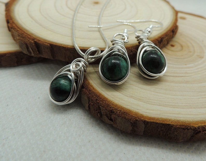 Puede incluir: Un conjunto de joyer&iacute;a que consta de un colgante en una cadena de tono plateado y dos pendientes a juego. Cada pieza presenta una piedra redonda de color verde oscuro y negro veteado, envuelta en un alambre plateado con dise&ntilde;o en espiral.