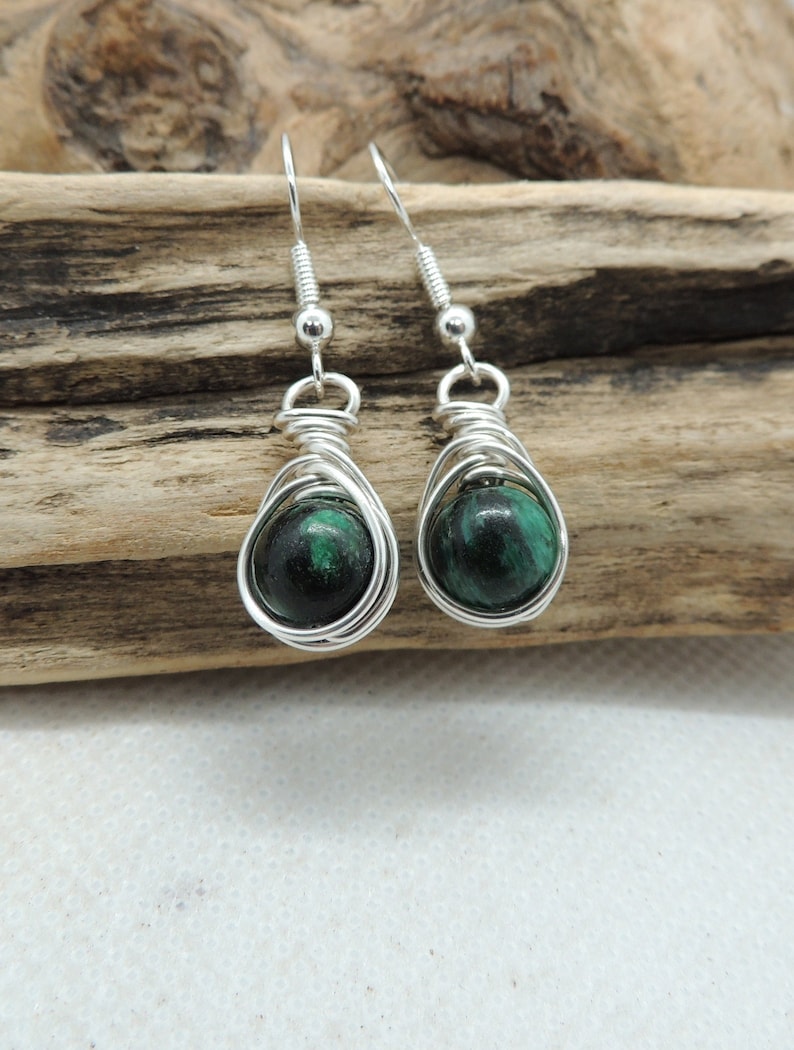 Puede incluir: Un par de pendientes colgantes con cuentas redondas de piedra veteada en verde oscuro y negro, envueltas en alambre de tono plateado. Los pendientes cuelgan de ganchos de plata y muestran un acabado pulido en las piedras esf&eacute;ricas.