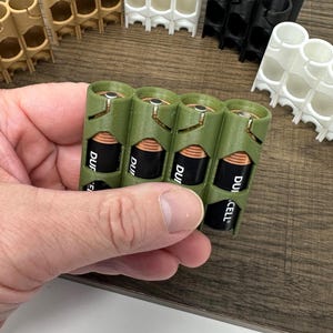 May include: Various 3D-printed battery holders in tan, green, black, and white. The holders are designed to store AA batteries. A hand holds a green holder with four AA batteries.