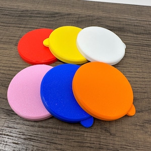 May include: A collection of seven colorful, round plastic lids in red, yellow, white, pink, blue, and orange. Each lid has a small tab on the side. The lids are arranged on a wooden surface.