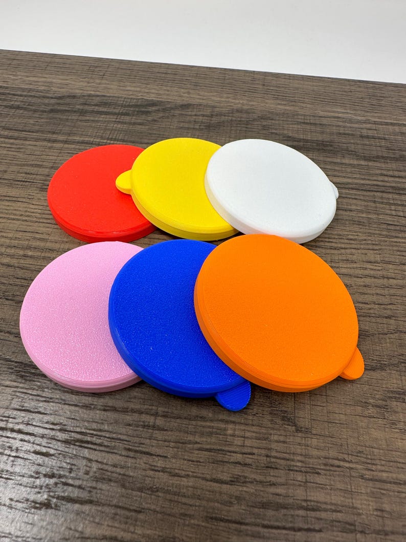 May include: A collection of seven colorful, round plastic lids in red, yellow, white, pink, blue, and orange. Each lid has a small tab on the side. The lids are arranged on a wooden surface.