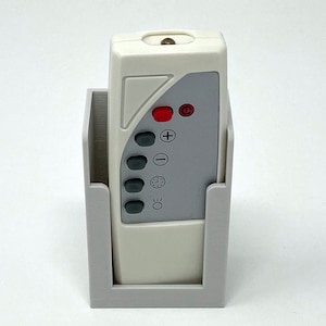 Puede incluir: Un mando a distancia blanco con una cara gris y varios botones, incluyendo un botón rojo, un signo más, un signo menos, un icono de reloj y un icono de bombilla. El mando a distancia está en un soporte de plástico blanco.