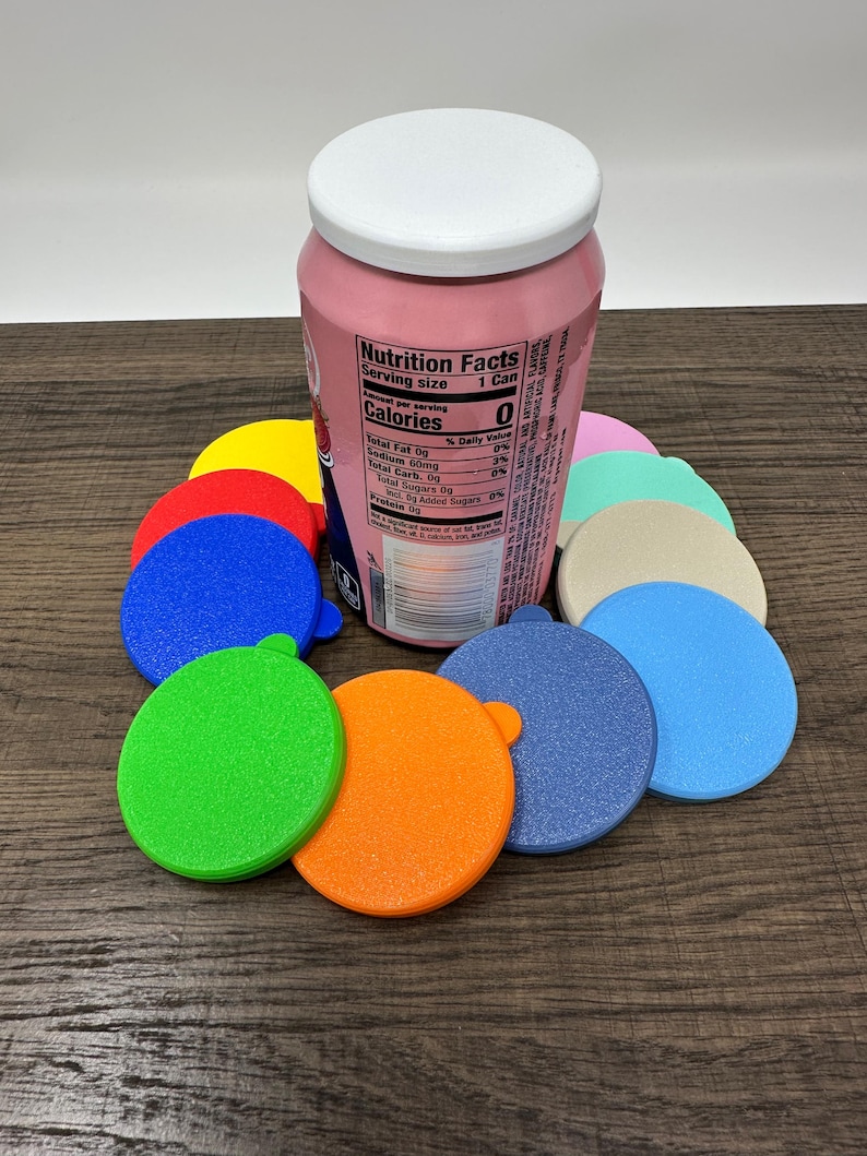 May include: A pink can with a white lid is surrounded by colorful, circular can covers in various colors, including yellow, blue, green, and orange. The can has a nutrition facts label. The covers have a small tab for easy removal.
