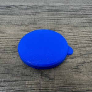 May include: A bright blue, round lid with a small tab for easy lifting. The lid is made of a textured material and is set against a wooden surface.