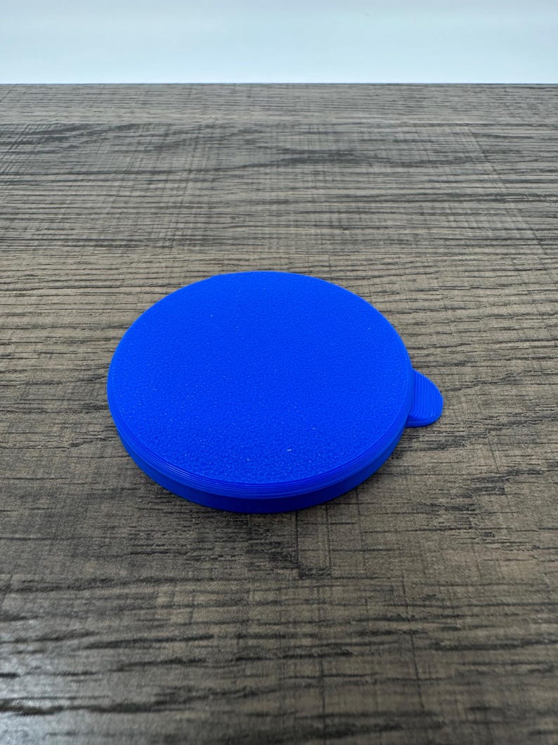 May include: A bright blue, round lid with a small tab for easy lifting. The lid is made of a textured material and is set against a wooden surface.