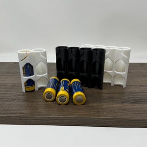May include: A collection of battery holders in black and white, with three yellow and blue batteries. The holders are designed to store and organize batteries. The image is set on a brown wooden surface.