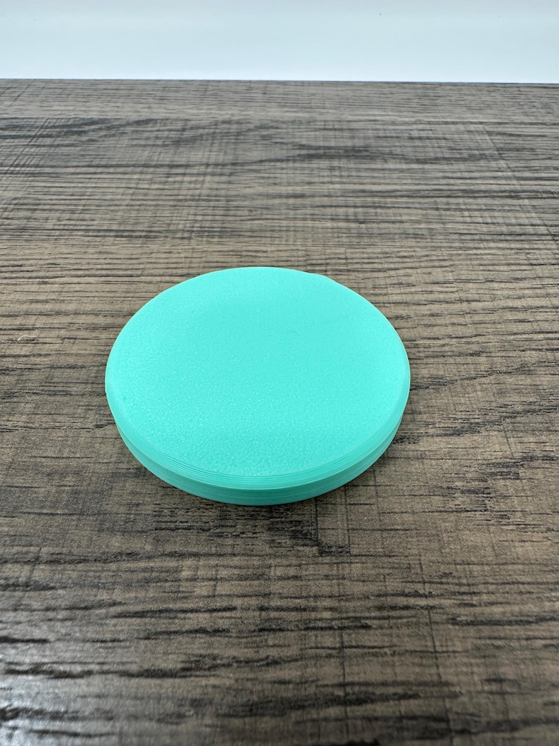May include: A round, teal-colored object with a slightly textured surface. The object is circular and appears to be made of a solid material. The item is sitting on a wooden surface.