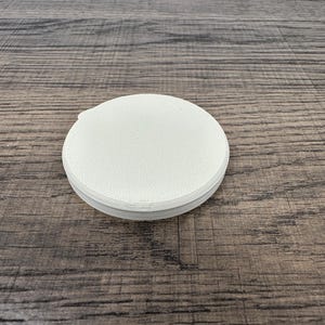 May include: A stack of white, round coasters with a textured surface. The coasters are made of a light-colored material and are stacked neatly on top of each other. The coasters are on a wooden surface.