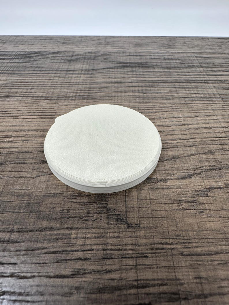 May include: A stack of white, round coasters with a textured surface. The coasters are made of a light-colored material and are stacked neatly on top of each other. The coasters are on a wooden surface.