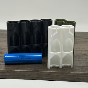 May include: A selection of 3D-printed battery holders in black, white, and olive green. A blue 3.7V Li-ion battery is placed in front of the holders. The holders are designed to securely hold cylindrical batteries, offering a practical storage solution.
