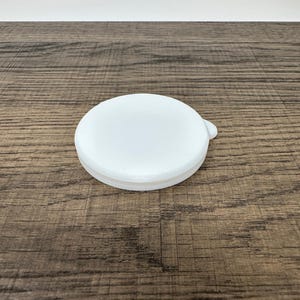May include: A white, round lid with a small tab on the side. The lid is made of a solid, opaque material and sits on a wooden surface. The lid is a simple, functional design.
