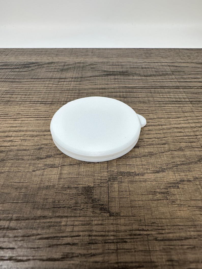 May include: A white, round lid with a small tab on the side. The lid is made of a solid, opaque material and sits on a wooden surface. The lid is a simple, functional design.