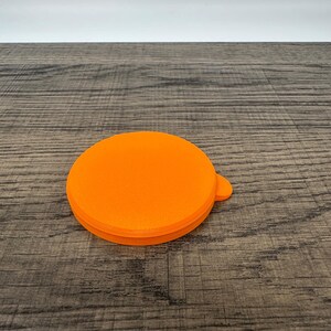 May include: Bright orange, circular lid with a small tab. The lid is made of a solid material and sits on a wooden surface. The lid's design is simple and functional, with a smooth, flat top and a slightly textured edge.