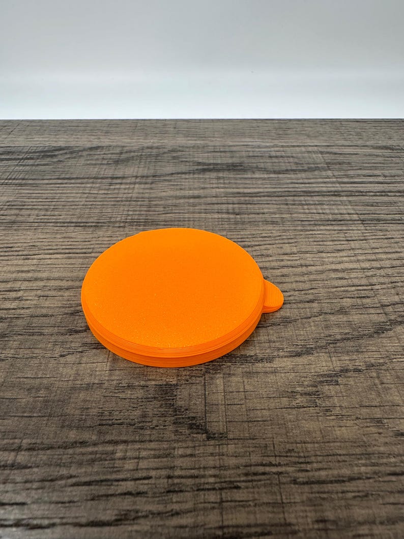 May include: Bright orange, circular lid with a small tab. The lid is made of a solid material and sits on a wooden surface. The lid's design is simple and functional, with a smooth, flat top and a slightly textured edge.