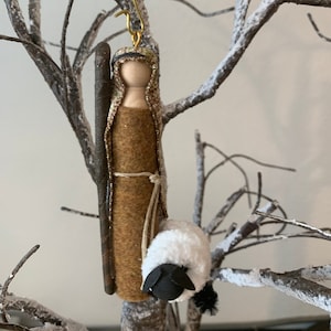 May include: A brown felt Christmas ornament depicting a shepherd with a white sheep. The shepherd is made of wood and has a brown felt robe. The ornament has a gold wire hanger.