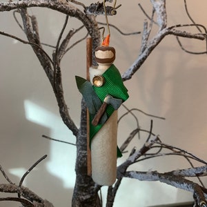 May include: A handmade felt Christmas ornament depicting a person with a green cloak, an axe, and a wooden staff. The ornament has a gold button and a brown beard.