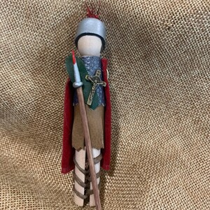 Könnte beinhalten: Eine Holzpuppe in Form eines Spielsteins, die als mittelalterlicher Ritter verkleidet ist. Die Puppe trägt einen roten Umhang, einen silbernen Helm und eine grün-silberne Tunika. Die Puppe hält eine braune Lanze mit einer roten Spitze.