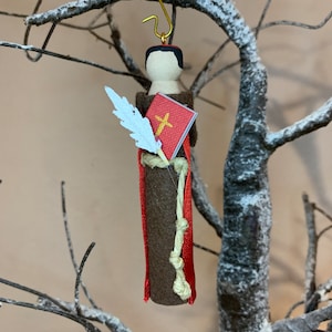 May include: A felt ornament depicting a saint holding a book and a quill. The saint is wearing a brown robe with a red sash and a white feather quill. The book is red with a gold cross on the cover.