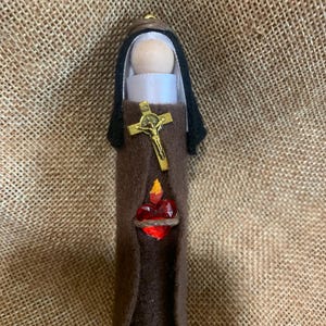 Könnte beinhalten: Eine braune Filzpuppe mit einem weißen Kopf und schwarzen Haaren. Die Puppe trägt ein weißes Gewand und eine braune Robe. Die Puppe hat ein goldenes Kreuz und ein rotes Herz mit einer Flamme oben drauf.
