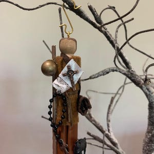 Puede incluir: Un adorno de madera que representa a un peregrino con una túnica marrón, una cruz, un rosario y un perro pequeño. El adorno está colgado de una rama.