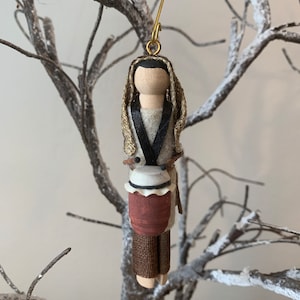 May include: A wooden ornament depicting a person with a brown and white drum. The person is wearing a brown robe with a gold trim and a black sash. The ornament is hanging from a gold wire.