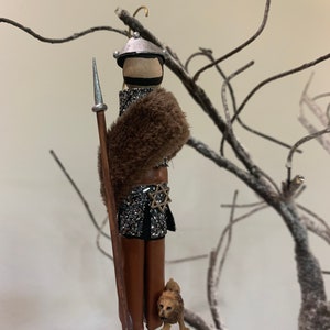 Peut inclure: Une figurine artisanale représentant un guerrier avec une lance, portant un casque argenté, une fourrure brune et une jupe de cotte de mailles argentée. La figurine tient une lance et a un petit chien brun à ses pieds.