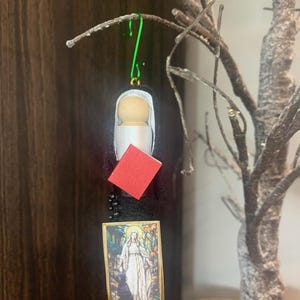 May include: A handmade religious ornament featuring a wooden peg doll dressed in white with a black veil. A small red book, a string of black beads, and a framed image of a saint are also included. The ornament hangs from a green wire hook.