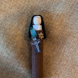 Puede incluir: Muñeca hecha a mano con cabeza de madera, pelo negro y cuerpo de fieltro marrón. La muñeca tiene un cuello blanco, una hoja verde, una estrella de David y una cruz con una representación de Jesús. La muñeca cuelga de un lazo dorado.