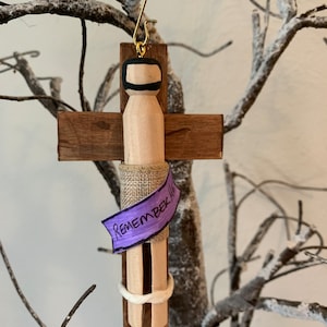 Może przedstawiać: Drewniana ozdoba w kształcie krzyża z drewnianą figurką na niej. Figurka jest owinięta w płótno i ma fioletową wstążkę z napisem "Pamiętaj o mnie".