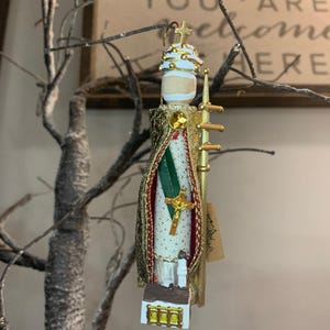 May include: A detailed Christmas ornament depicting a religious figure. The ornament features a gold robe, a white and green interior, and a small church at the bottom. A gold cross is visible on the front. The ornament is hanging from a tree branch.