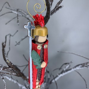 May include: A handmade Christmas ornament depicting a Roman soldier in red and gold armour, holding a silver cross and a green palm leaf. The ornament is made of wood and fabric and has a gold wire hanger.