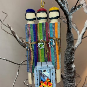 May include: Three wooden figures dressed in colorful striped robes, with blue, red, and yellow hats, stand in front of a small blue wooden structure with a fire inside. The figures are holding silver stars with a Star of David design.