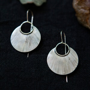 May include: A pair of silver earrings with a fan-shaped design. The earrings have a circular hoop at the top and hang from a silver hook.