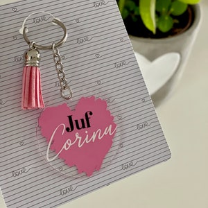 Op de afbeelding: Roze hartvormige sleutelhanger met de tekst "Juf Corina" erop. De sleutelhanger heeft een zilveren ketting en een roze kwast.
