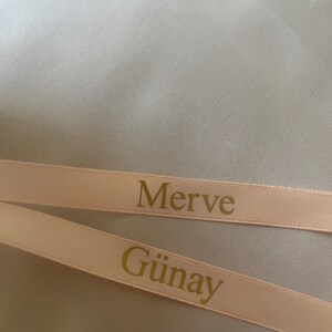 May include: Two light pink ribbons with gold text. The top ribbon reads "Merve" and the bottom ribbon reads "G&uuml;nay". The ribbons are set against a light gray background.