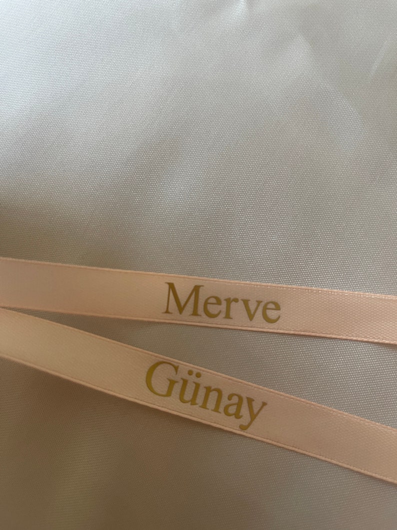 May include: Two light pink ribbons with gold text. The top ribbon reads "Merve" and the bottom ribbon reads "G&uuml;nay". The ribbons are set against a light gray background.