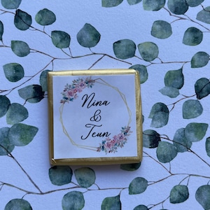 May include: A square chocolate candy wrapped in gold foil. The label features the names "Nina & Teun" in elegant script within a gold geometric frame, adorned with floral accents. The background is a white surface with a pattern of green eucalyptus leaves.