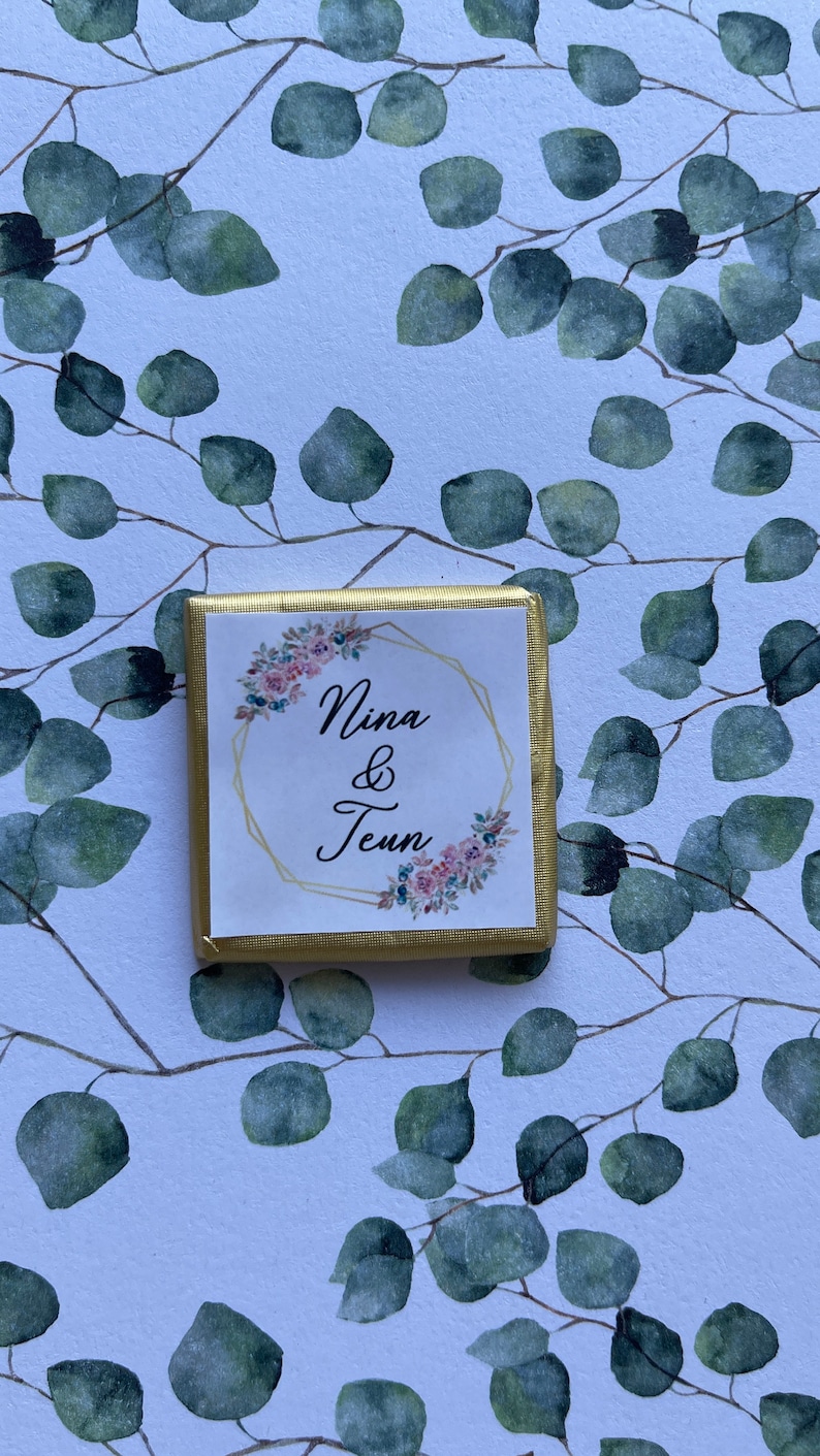 May include: A square chocolate candy wrapped in gold foil. The label features the names "Nina & Teun" in elegant script within a gold geometric frame, adorned with floral accents. The background is a white surface with a pattern of green eucalyptus leaves.