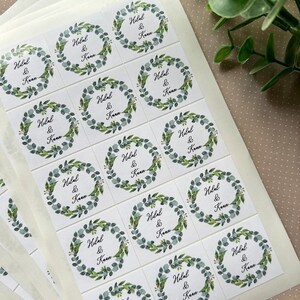May include: Square white stickers with a watercolor eucalyptus wreath design. Each sticker features the names "Hilal & Kaan" in a handwritten font. The stickers are arranged on a white backing sheet, suitable for wedding favors or event stationery.