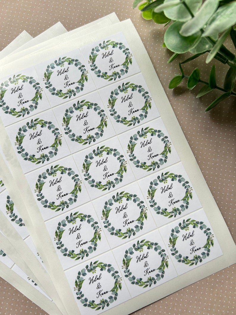 May include: Square white stickers with a watercolor eucalyptus wreath design. Each sticker features the names "Hilal & Kaan" in a handwritten font. The stickers are arranged on a white backing sheet, suitable for wedding favors or event stationery.