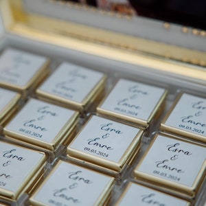 May include: A box of chocolates with gold foil wrappers. Each chocolate square has a white label with the names "Esra & Emre" and the date "09.03.2024" in elegant script. The box has a gold trim.