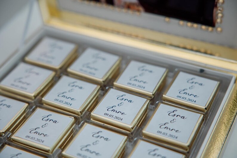 May include: A box of chocolates with gold foil wrappers. Each chocolate square has a white label with the names "Esra & Emre" and the date "09.03.2024" in elegant script. The box has a gold trim.